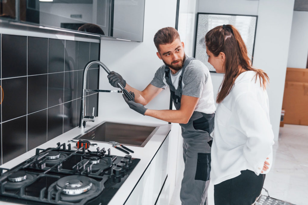 Young professional plumber in grey uniform shows results of work for housewife on the kitchen